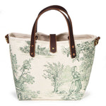 Front of the Butter and Pine Toile Mini Tote, a young woman with a basket beside a cottage and grapevines, with mahogany handles and button tab