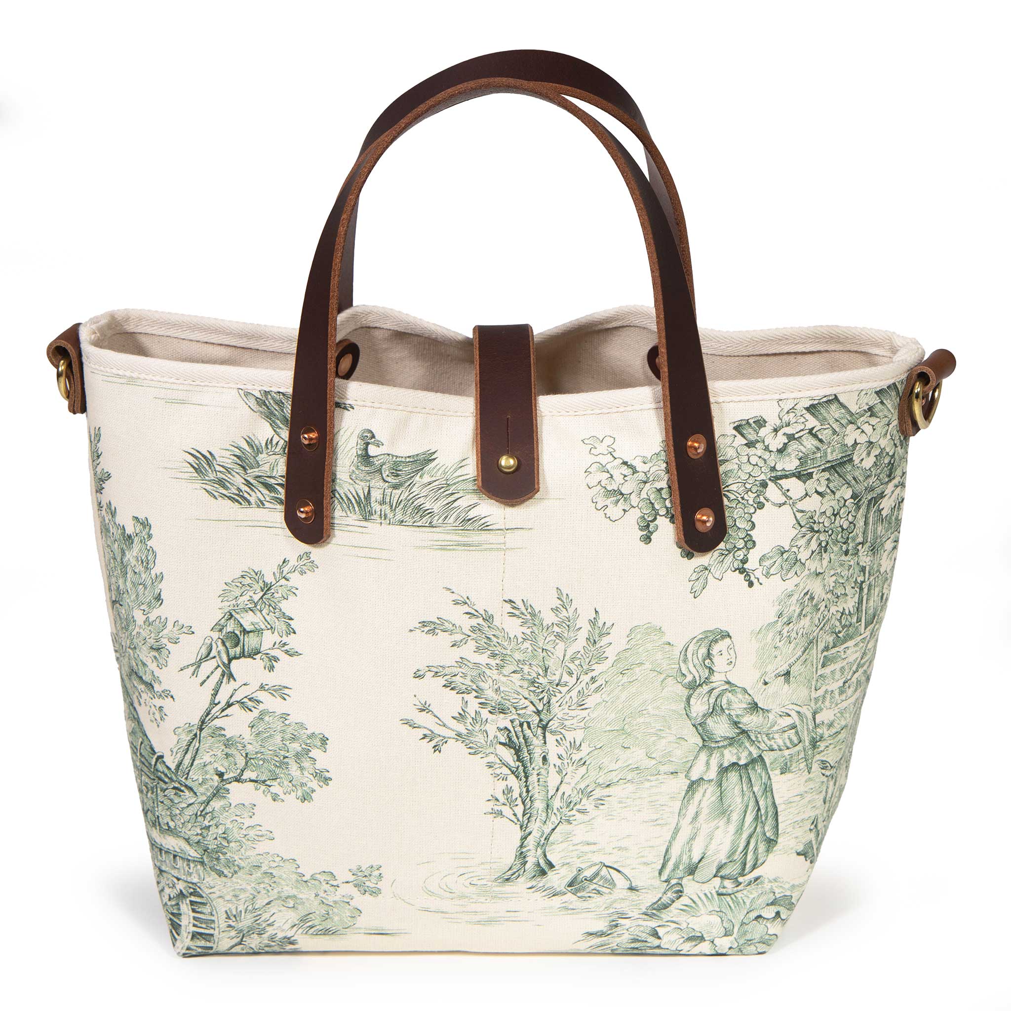 Front of the Butter and Pine Toile Mini Tote, a young woman with a basket beside a cottage and grapevines, with mahogany handles and button tab