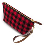 The Buffalo Check Travel Clutch, red and black buffalo check, shown with its detachable mahogany leather wristlet clipped to a brass D-ring