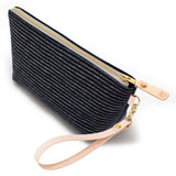 The Indigo Chalk Stripe Travel Clutch, white chalk stripes on indigo, shown with its detachable blonde leather wristlet clipped to a brass D-ring