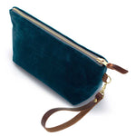 The Ocean Velvet Travel Clutch, ocean teal velvet, shown with its detachable mahogany leather wristlet clipped to a brass D-ring
