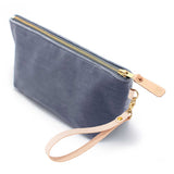The Cloudy Velvet Travel Clutch, blue-grey velvet, shown with its detachable blonde leather wristlet clipped to a brass D-ring