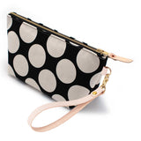 The Monster Dot Travel Clutch, oversized cream dots on black, shown with its detachable blonde leather wristlet clipped to a brass D-ring