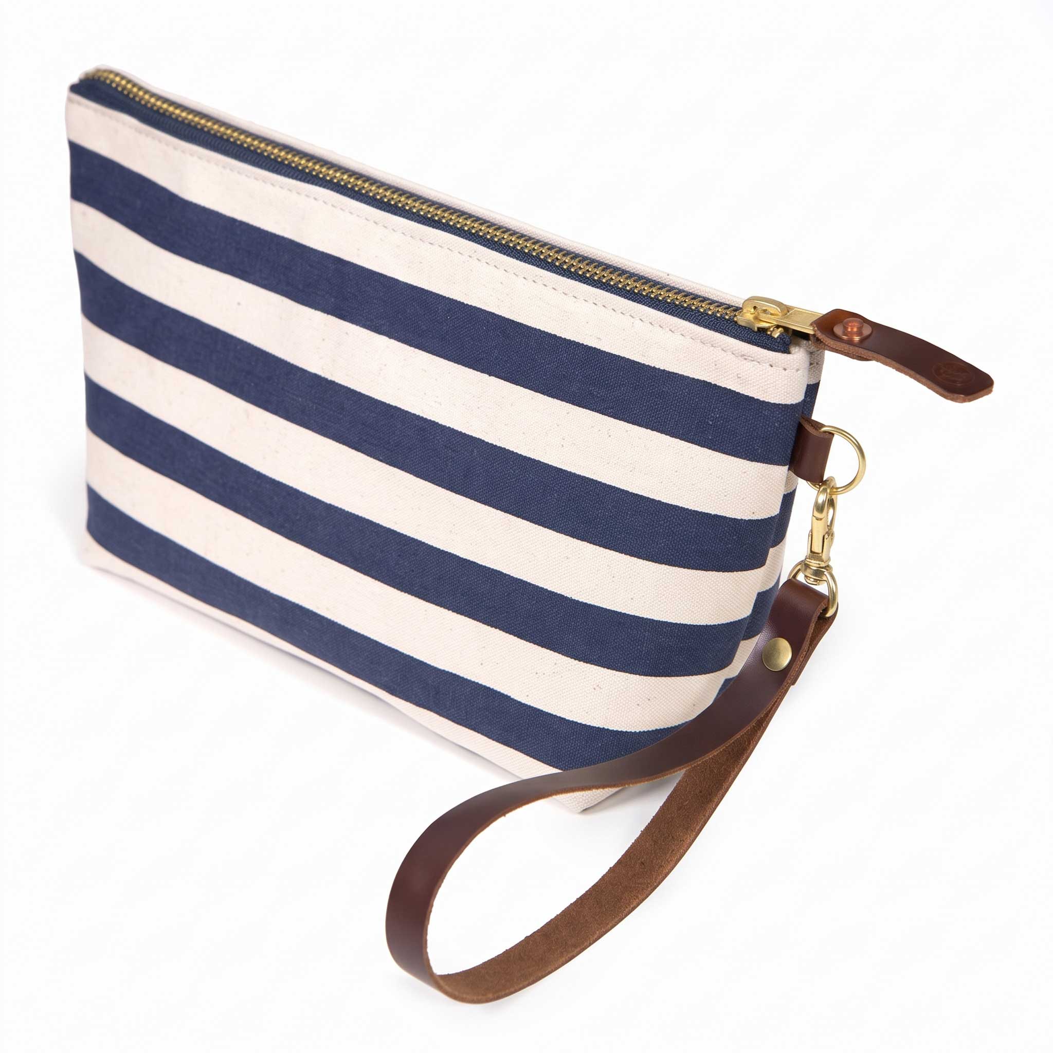 The Navy Cabana Stripe Travel Clutch, wide navy and white stripes, shown with its detachable mahogany leather wristlet clipped to a brass D-ring