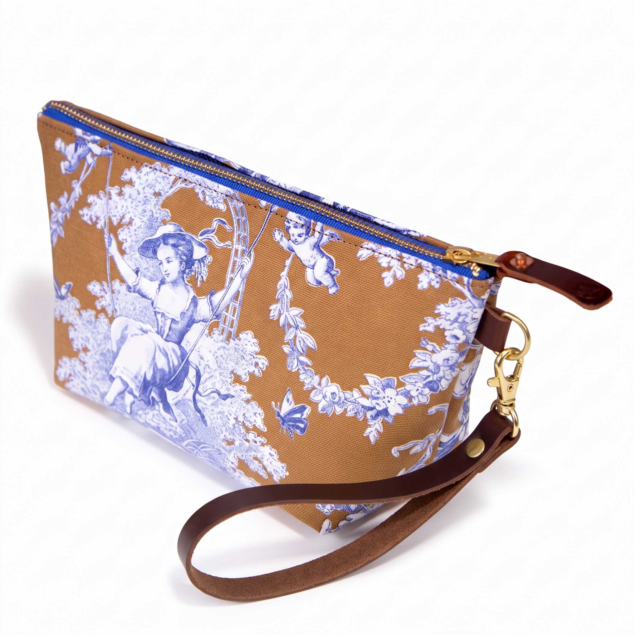 The Oak and Ink Toile Travel Clutch, blue toile scenes on oak brown, shown with its detachable mahogany leather wristlet clipped to a brass D-ring