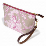 The Sand and Rose Toile Travel Clutch, rose-pink toile scenes on sand, shown with its detachable mahogany leather wristlet clipped to a brass D-ring