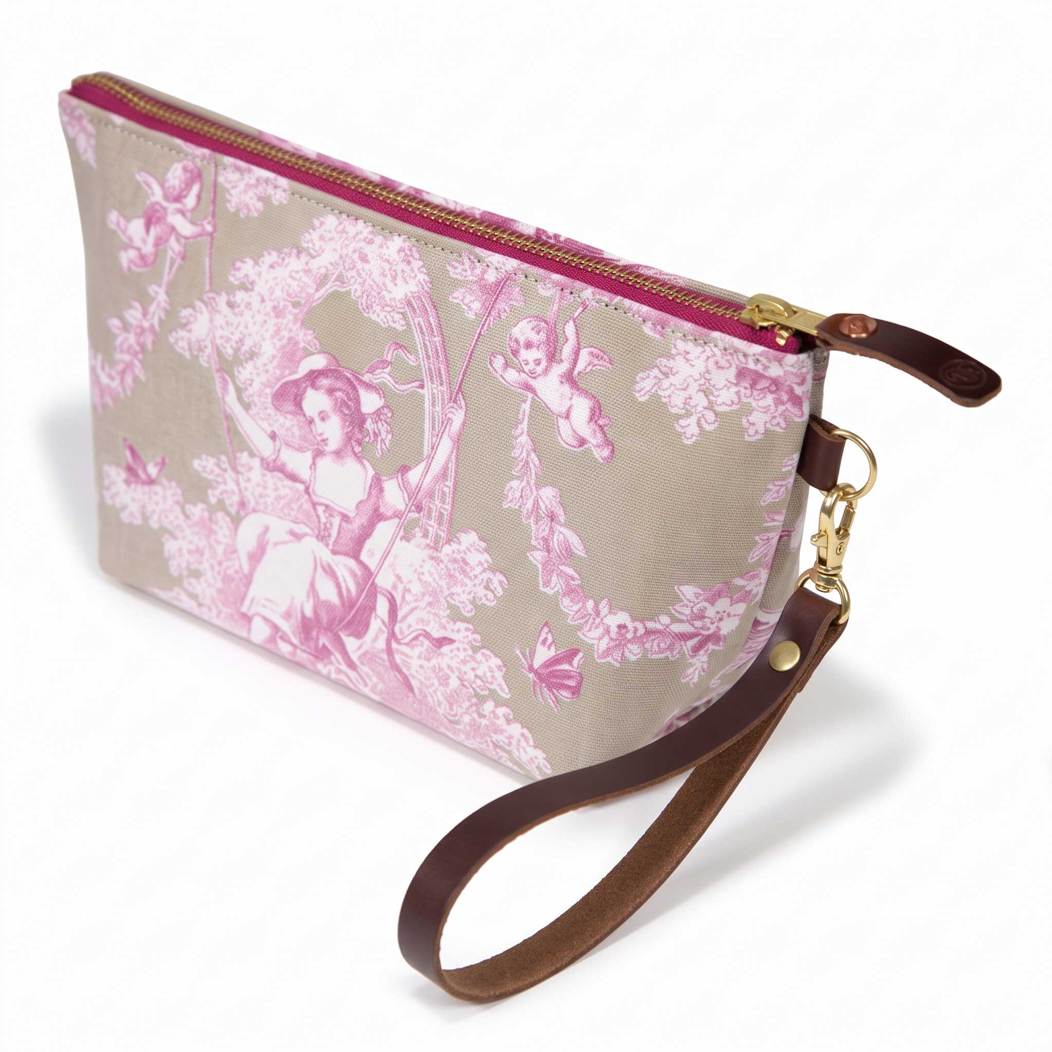 The Sand and Rose Toile Travel Clutch, rose-pink toile scenes on sand, shown with its detachable mahogany leather wristlet clipped to a brass D-ring