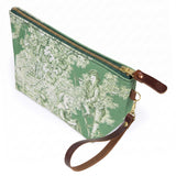 The Emerald Toile Travel Clutch, cream toile scenes on emerald green, shown with its detachable mahogany leather wristlet clipped to a brass D-ring