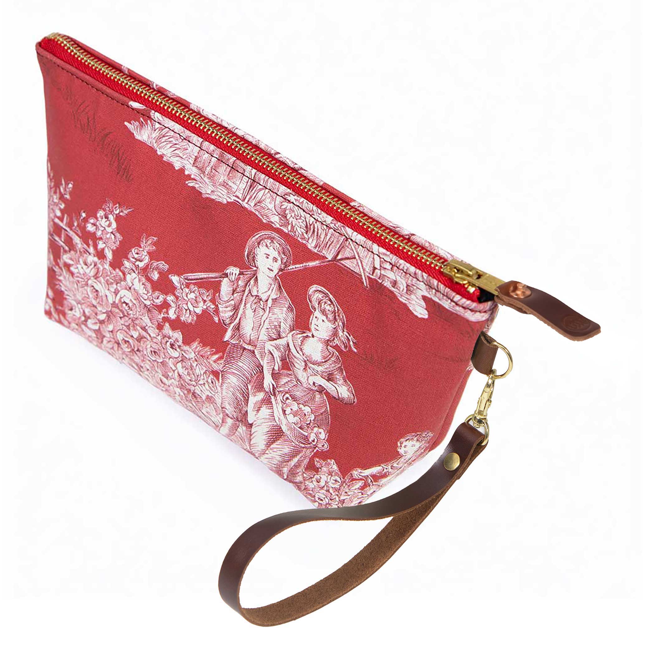 The Scarlet Toile Travel Clutch, white toile scenes on scarlet, shown with its detachable mahogany leather wristlet clipped to a brass D-ring
