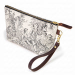 The Cream & Coal Toile Travel Clutch, charcoal toile scenes on cream, shown with its detachable mahogany leather wristlet clipped to a brass D-ring