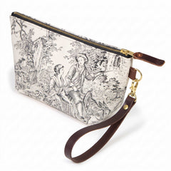 The Cream & Coal Toile Travel Clutch, charcoal toile scenes on cream, shown with its detachable mahogany leather wristlet clipped to a brass D-ring