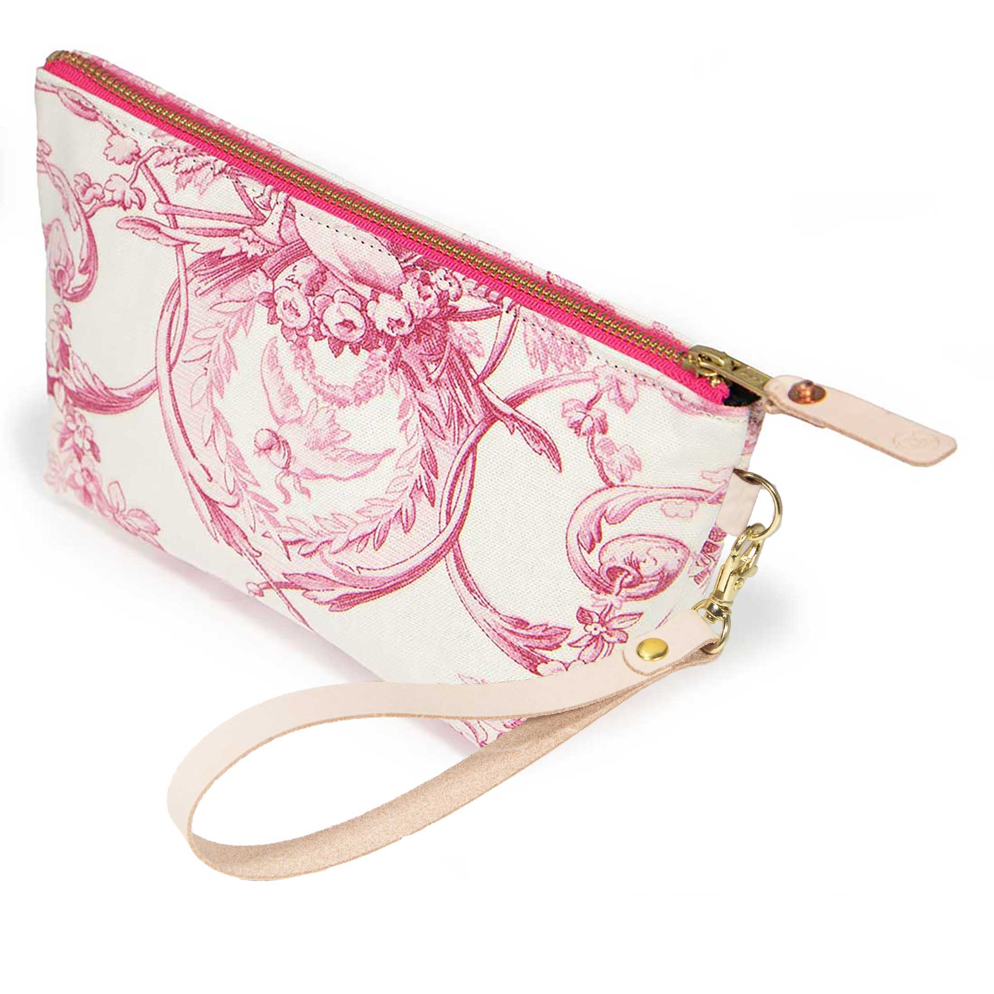 The French Rose Toile Travel Clutch, rose-pink toile scenes on cream, shown with its detachable blonde leather wristlet clipped to a brass D-ring