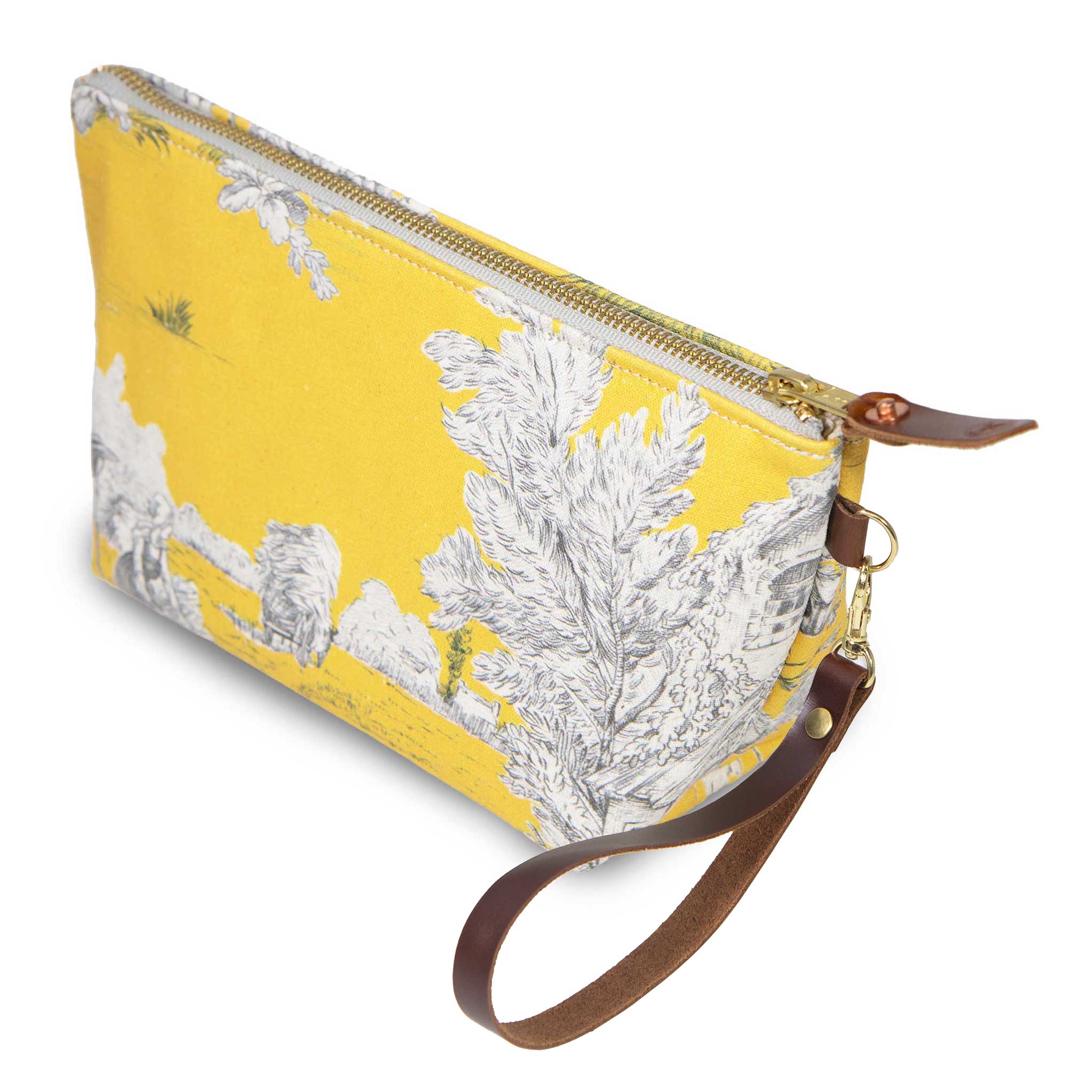 The Sunshine Toile Travel Clutch, white toile scenes on yellow, shown with its detachable mahogany leather wristlet clipped to a brass D-ring