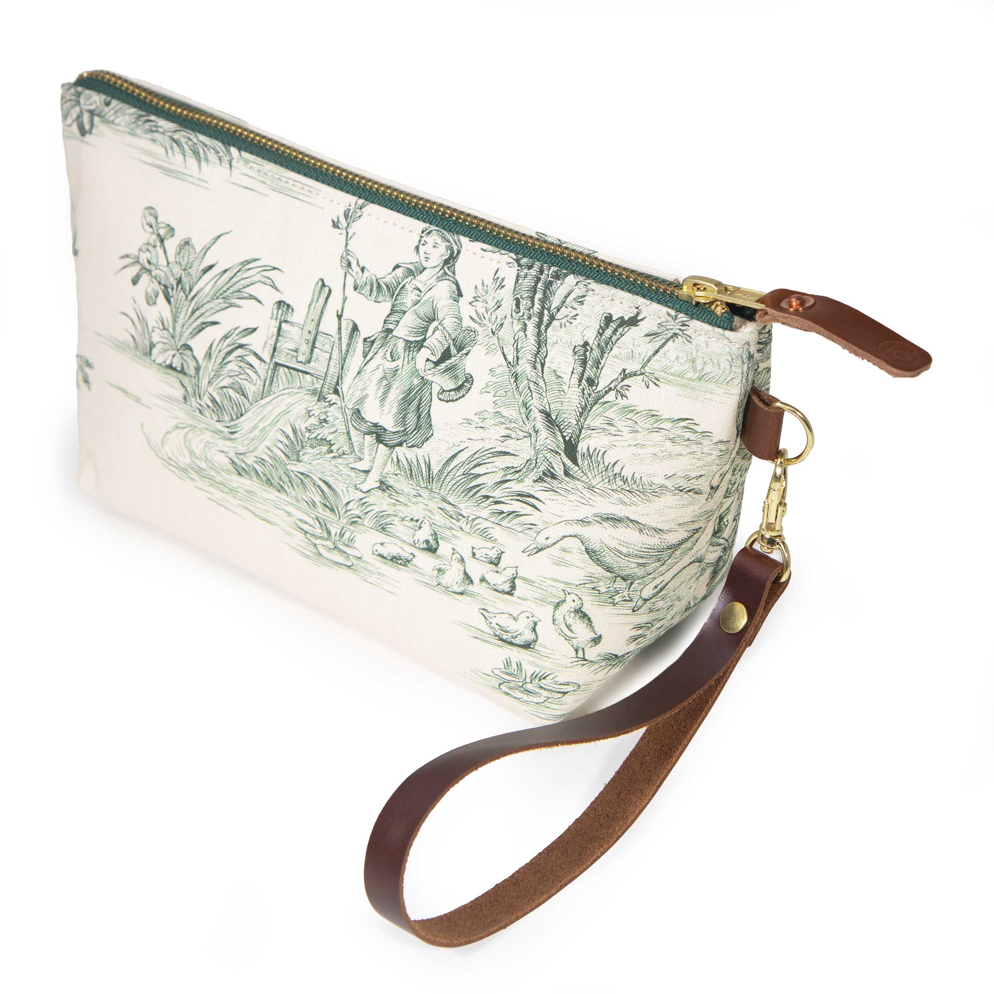 Butter and Pine Toile Travel Clutch with its detachable leather wristlet clipped to a brass D-ring, showing a young woman at the water's edge in pine green toile